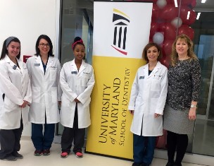 Sheryl Syme and three dental hygiene students in white coats standing by a banner for the University of Maryland School of Dentistry