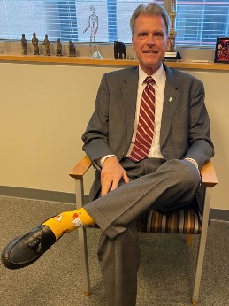 Dean Reynolds sits in a chair, showing off a pair of colorful socks